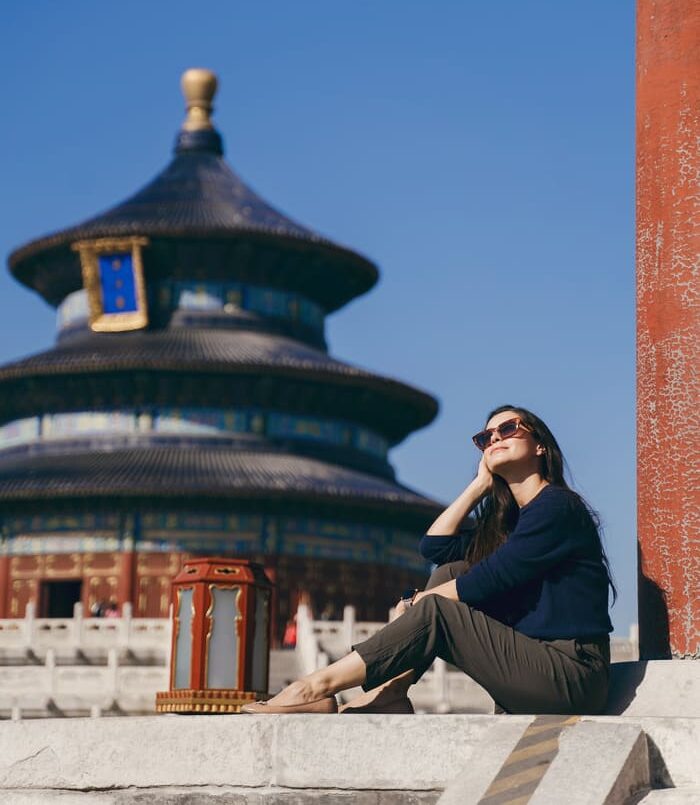 Una mujer sentada junto al Templo del Cielo, bajo un cielo despejado, mira hacia el sol con gafas de sol en Beijing.