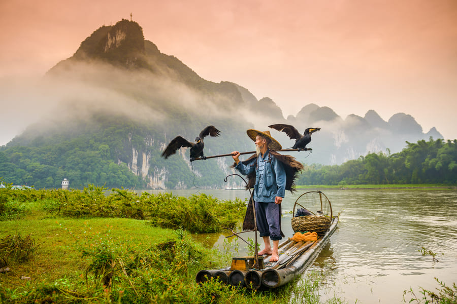 Un pescador de pie sobre una balsa de bambú sostiene dos cormoranes junto a un río, con montañas cubiertas de niebla al fondo